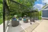 Charming outdoor space featuring modern pergolas, cozy wicker chairs with blue cushions, and built-in BBQ grills against a lush green backdrop.