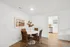 Bright dining area with elegant wicker chairs around a round white table, complemented by modern wall art and a glimpse into a sleek kitchen with stainless steel appliances.