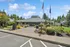 Inviting apartment complex entrance with vibrant landscaping, featuring mature trees, colorful flowers, and a well-maintained path under a clear blue sky.