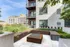 Modern apartment exterior featuring a stylish patio with lush greenery, contemporary seating, and a chic firepit under a beautiful blue sky.