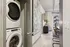 Modern apartment corridor showcasing sleek laundry area with stacked washer and dryer, leading to bright living space with contemporary decor and panoramic window view.