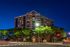 Elegant apartment building exterior at night, illuminated with warm lighting, surrounded by lush greenery and city vibes.
