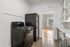 Modern kitchen with sleek black appliances, bright cabinetry, and a view into the sunlit living room featuring large windows and hardwood flooring.