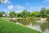 Idyllic apartment complex nestled by a serene pond with fountains, surrounded by lush greenery and immaculate lawns under a clear blue sky.