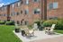 Charming brick apartment exterior with a sunlit patio featuring cozy Adirondack chairs, surrounded by lush greenery.
