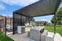 Modern outdoor barbecue area with a shaded pergola, sleek grill, and concrete picnic table, set against a vibrant green lawn and playground backdrop.
