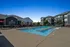 Modern apartment complex featuring a sparkling outdoor swimming pool with stylish poolside loungers and umbrellas, set against a clear blue sky.