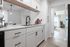 Modern kitchen with sleek white cabinets, elegant countertops, and a stylish faucet, opening into a bright living space with natural light.