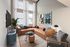 Modern living room with high ceilings, featuring a luxurious brown sectional sofa, stylish artwork, and ample natural light from large windows.