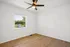 Bright, modern apartment room with sleek wood flooring, a ceiling fan, and a large window featuring a pleasant view of greenery outside.