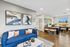 Modern apartment interior with a chic blue sofa, abstract wall art, and a bright workspace featuring wooden chairs and large windows.