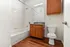 Modern bathroom with elegant wooden cabinetry, sleek countertop, and a cozy bathtub. Features stylish wood flooring and pristine white tiles, creating a fresh and inviting space.