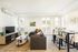 Modern, bright living and dining area featuring a cozy l-shaped sofa, flat-screen TV, wooden furniture, and large windows with scenic views.