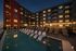 Elegant apartment complex with a beautifully illuminated poolside lounge, numerous cozy deck chairs, and string lights creating a warm, inviting evening ambiance.