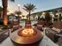 Luxurious outdoor lounge area with a cozy fire pit surrounded by stylish wicker seating, palm trees, and a serene pool backdrop at sunset.
