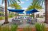 Luxurious poolside oasis with elegant patio tables, shaded by vibrant blue umbrellas, surrounded by lush palm trees and modern apartments.