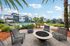 Luxurious apartment complex featuring a resort-style pool, modern fire pit seating area, lush landscaping, and shaded pergolas under sunny skies.