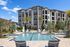 Modern apartment complex with a refreshing pool, surrounded by lush palm trees and lounging chairs, under a clear blue sky.