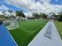 Vibrant outdoor sports field with lush green artificial turf, surrounded by a scenic neighborhood and blue skies, perfect for recreational activities.