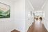 Bright and inviting hallway with elegant wood flooring, white paneled walls, and a glimpse of a cozy living room adorned with large windows, modern art, and stylish decor.