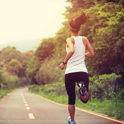 A woman in a white shirt jogging on a paved trail in a wooded park setting.